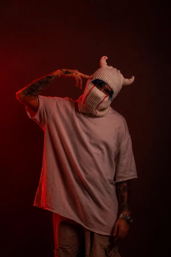 SEVEN77 con pasamontañas blanco de cuernos y camiseta oversize, bajo luz roja sobre fondo oscuro